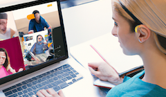 A woman sits at her computer looking at the screen which has four people in indivudal squares of a video conference call.