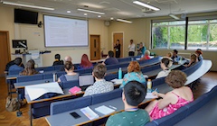 Students sit in a lecture-theater style classroom while a professor gives a presentation at the front of the room.
