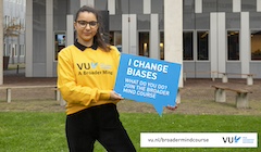 A student wearing a Vrije Universiteit Amsterdam sweatshirt holds a sign saying "I Change Business."